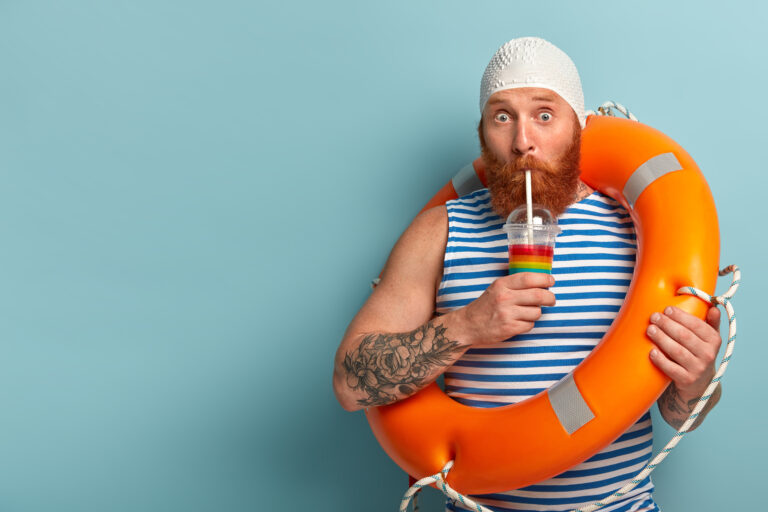 Embarrassed holiday maker drinks cold summer cocktail, spends free time at beach, wears swimcap sailor t shirt, swims with lifebuoy, has surprised expression, models over blue wall with free space